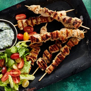 chicken souvlaki skewers surrounded by a bowl of tzatziki, cherry tomatoes, and chopped romaine on a black platter on a dark green background