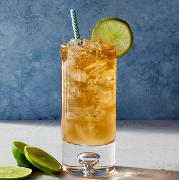 glass of a dark and stormy cocktail with a paper straw and a lime sitting on a stone surface with a marbled blue background